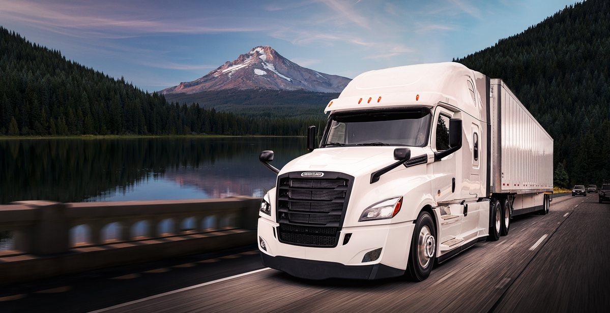 White semi tractor with dry van trailer running past a lake with a snow capped mountain behind it